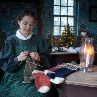 A woman in 1830s dress knits a pair of stocking by candlelight