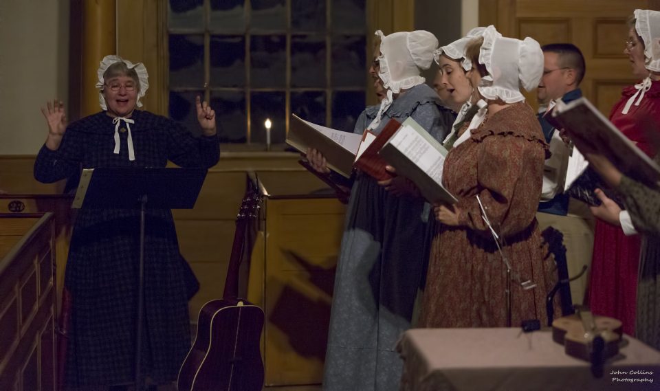 A choir dressed in 1830s attire performs inside a meetinghouse